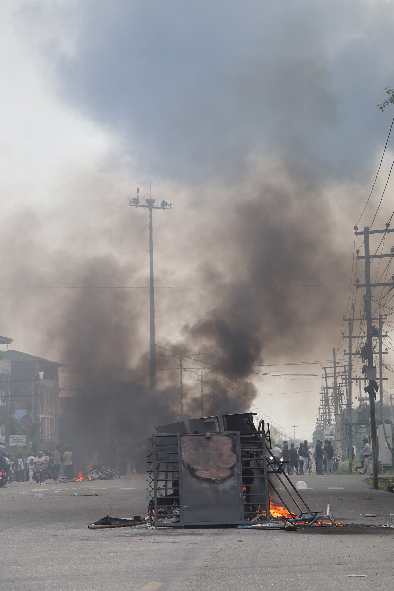 Black smoke rising from a burning barricade in the middle of a street in Chitwan, Nepal, with a crowd of protesters visible in the distance, September 9, 2025